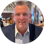 Perry Belcher, a man with glasses and short hair, smiles at the camera while wearing a suit in a bustling restaurant filled with lively patrons in the background.